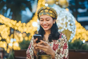 A cheerful woman in traditional clothing using her smartphone during a festive moment, showing how easily the PHCHARMS app fits into everyday life.